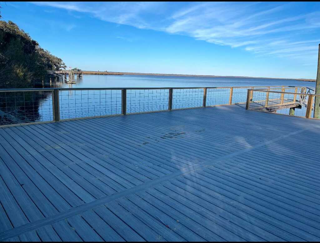 A spacious wooden deck overlooks a calm lake under a bright blue sky. Greenery edges the water, creating a serene and peaceful atmosphere.