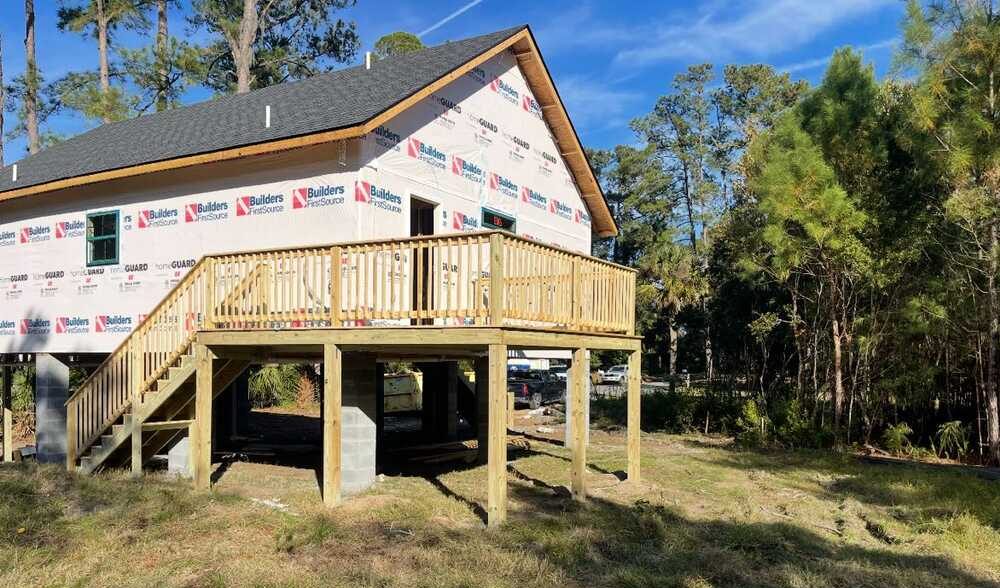 A partially constructed house wrapped in protective material with a wooden deck and stairs. It's elevated on stilts, surrounded by trees under a clear blue sky.