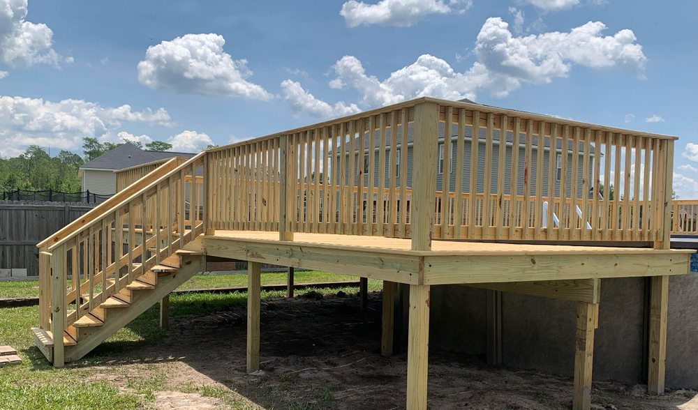 Elevated wooden deck with railings and a staircase, set against a blue sky with clouds. The structure sits in a grassy backyard, suggesting a clean, fresh atmosphere.