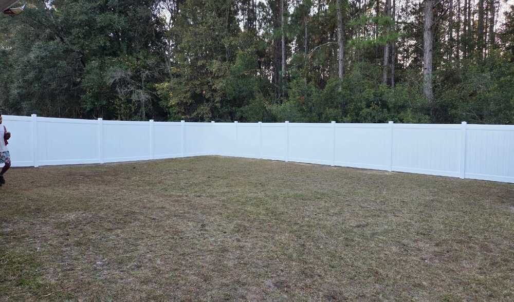 A white vinyl fence encloses a grassy yard area, bordered by dense green trees. The scene is quiet and serene, with a clear, spacious sky above.