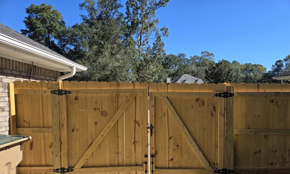 Wooden double gate with diagonal cross-bracing stands between two brick walls. Sunlight creates sharp shadows. Trees and blue sky in the background.