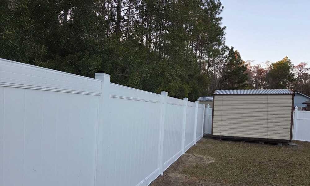 A tall white fence runs through a backyard, bordering dense green trees. A beige shed with a metal roof stands on the right, creating a serene, suburban scene.